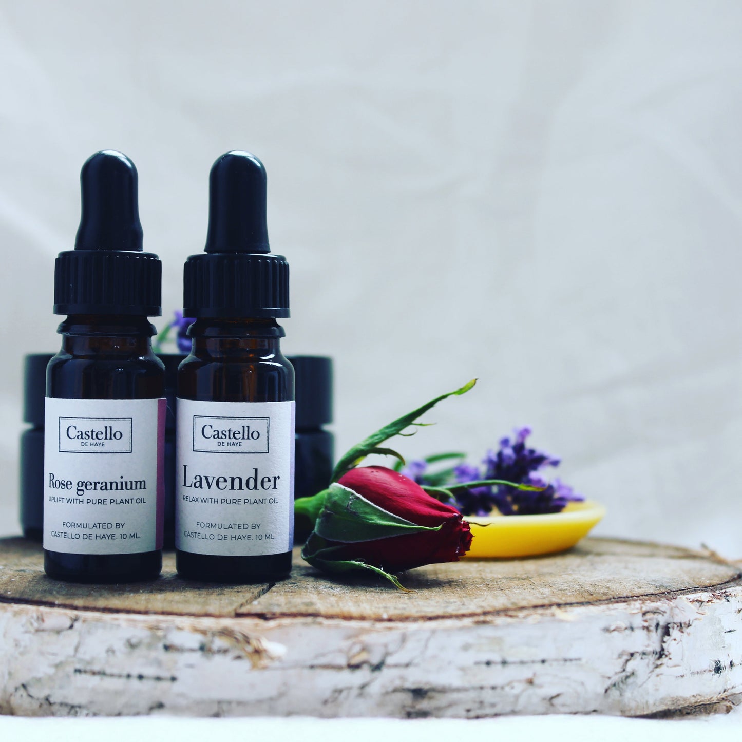10ml bottles of rose geranium essential oil and lavender essential oil on wooden tray with a rose flower and lavender in the background